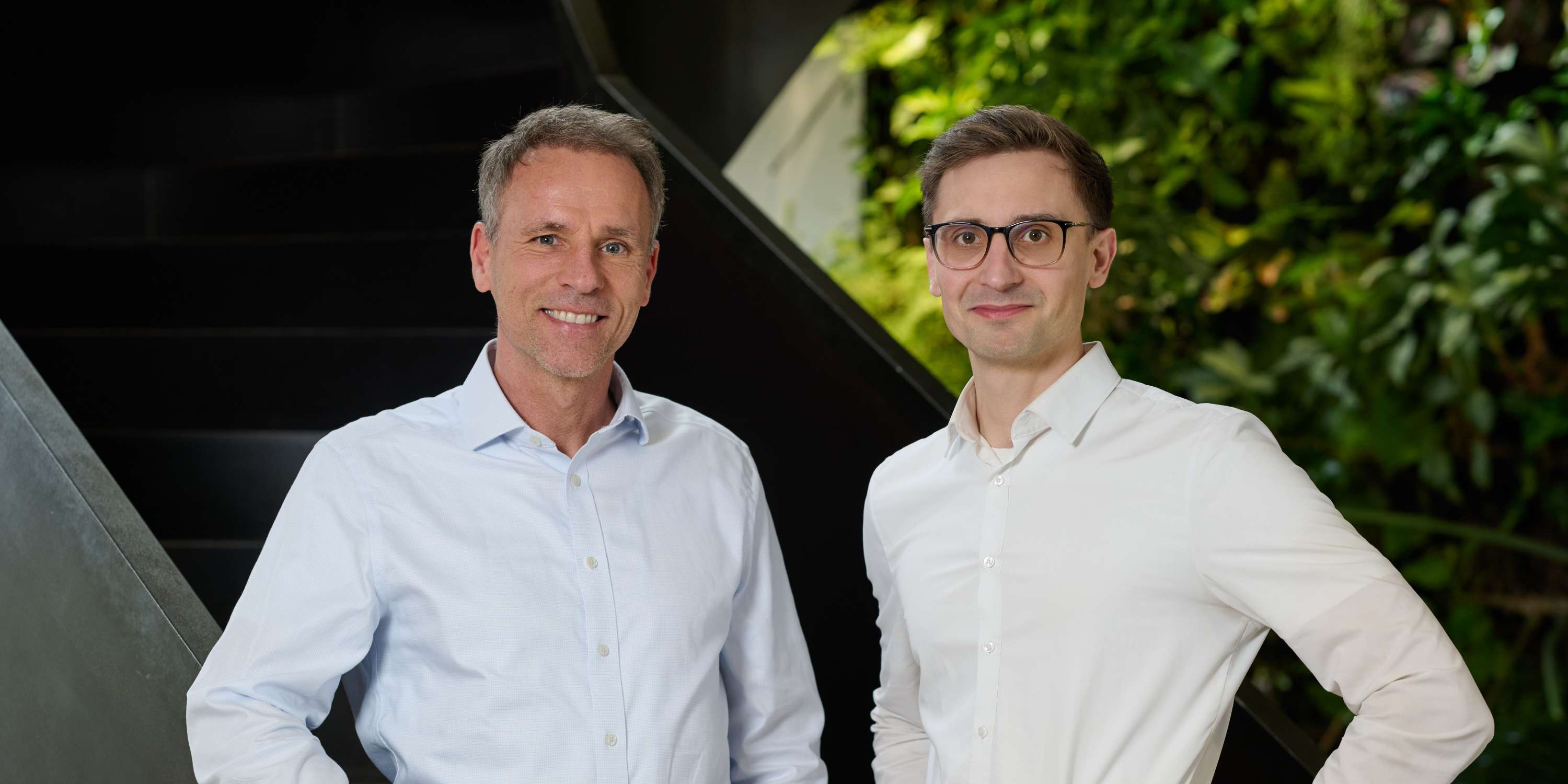 Ein Porträtfoto zweier Männer, die nebeneinander auf einer Treppe posieren. Links steht Stefan Henze, ein lächelnder Mann mit grauem, kurzem Haar in einem hellblauen gestreiften Hemd und grauen Hosen, die Hände auf dem Geländer. Rechts steht Eric Yeung-Schüürmann, ein junger Mann mit Brille in einem weißen Hemd und dunklen Hosen, die Hände in den Hüften. Im Hintergrund ist eine grüne Pflanzenwand zu sehen.