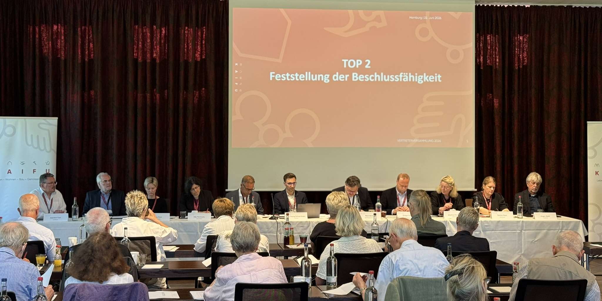 KAIFU-Vertreterversammlung: Ein Konferenzraum mit vielen Menschen an Tischen, die einer Leinwand zugewandt sind, auf der "65. ORDENTLICHE VERTRETERVERSAMMLUNG" steht.