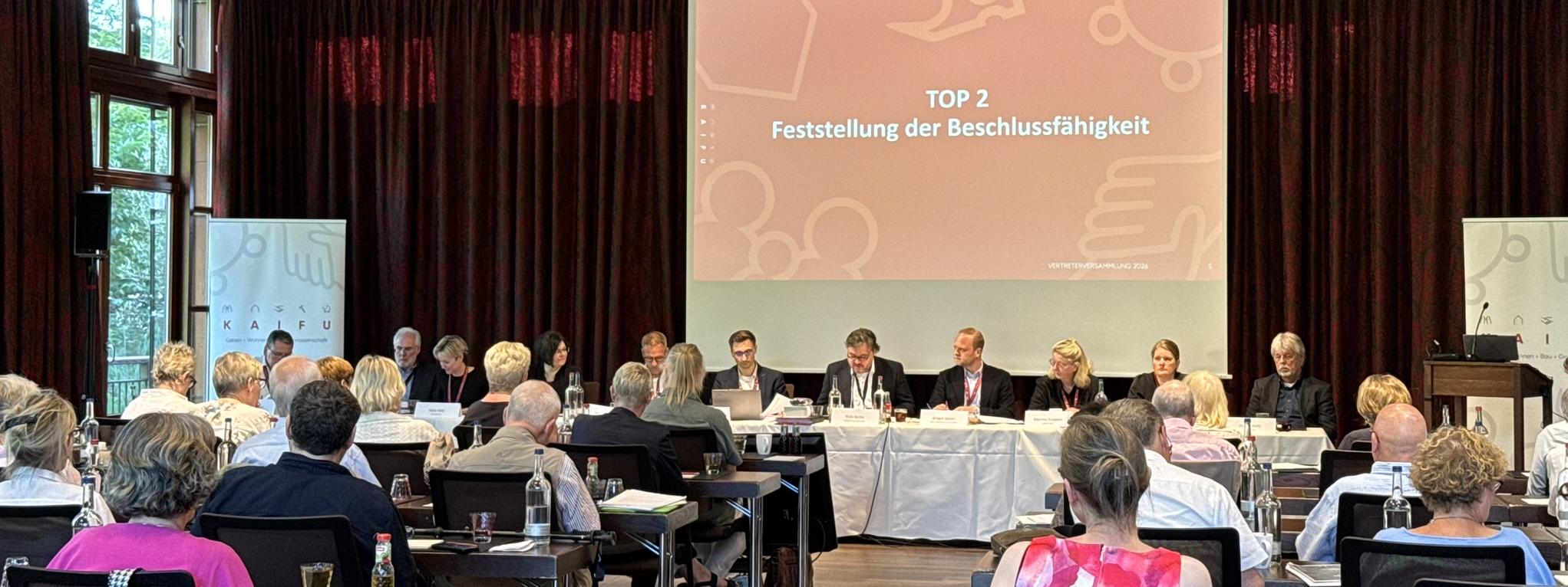KAIFU-Vertreterversammlung: Ein Konferenzraum mit vielen Menschen an Tischen, die einer Leinwand zugewandt sind, auf der "65. ORDENTLICHE VERTRETERVERSAMMLUNG" steht.